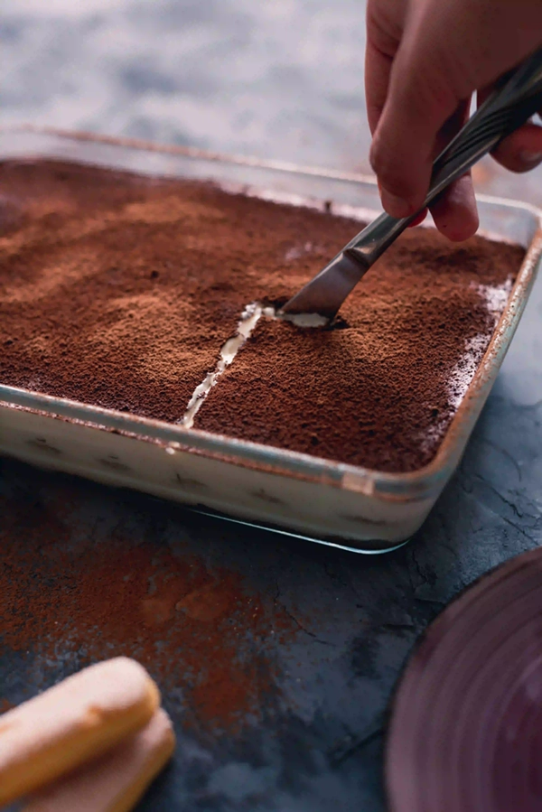 En person skär upp en hel tiramisu i fyrkantiga bitar för servering.